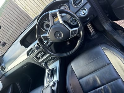 The scene depicts the interior of a Mercedes-Benz passenger vehicle, observed from the front passenger compartment, facing the driver's area and front windshield. The setting is Montrose, United Kingdom. The vehicle's interior is characterized by black leather seating surfaces, a black multi-function steering wheel emblazoned with the Mercedes-Benz logo, and a dashboard featuring a horizontal silver trim panel. An automatic transmission gear selector with a black leather gaiter and chrome elements is situated on the center console, alongside climate controls and a dark central display unit. The driver's footwell contains two silver, perforated metal pedals. Exterior visibility through the windshield reveals a light-toned wooden fence composed of horizontal slats, indicating an outdoor environment. Direct natural sunlight is evident, illuminating various interior surfaces and creating reflections. No occupants are present within the vehicle.