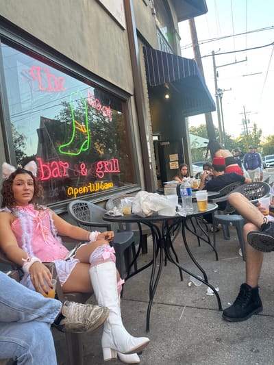 An outdoor scene is depicted on the sidewalk patio of "The Boot bar & grill" in New Orleans, United States. The establishment is identifiable by prominent pink and green neon signs in the large front window, displaying "The Boot", a boot graphic, "bar & grill", and "opentill6am".
In the immediate foreground, a female with curly brown hair is seated on a dark grey plastic patio chair. She wears a pink and white "cat girl" costume, consisting of a pink corset with white fur trim and bows, a white ruffled mini-skirt, white knee-high boots with pink bows, white wrist cuffs, and a white cat-ear headband. She holds a clear plastic cup containing an amber liquid and is looking directly at the viewer. To her left, the legs of another individual in blue jeans and dirty white sneakers are partially visible.
A round, dark metal patio table stands between the foreground subjects and the building. It is cluttered with multiple clear plastic cups containing amber beverages, a plastic water bottle, and crumpled white paper/plastic debris. Several dark grey plastic chairs surround this table and another similar table further back.
At the rear table, several individuals are seated. One person, with their back partially to the camera, wears a red fez and a dark shirt. Another female with dark hair is seated opposite them. In the background, a woman with long dark hair, wearing a red top and patterned skirt, is seen standing near the tables. Beyond the patio, a bustling street scene includes utility poles, power lines, and distant pedestrians and vehicles. The lighting suggests late afternoon or early evening.