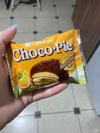 An eye-level shot captures a light-skinned hand, possibly female, holding a single-serving package of "ORION Choco.Pie" with a prominent orange flavor. The hand's thumb and index finger, adorned with iridescent light blue nail polish, cradle the bottom and top of the package respectively.

The snack's packaging is predominantly bright orange and yellow. The brand name "ORION" and "Choco.Pie" are clearly visible, with "Choco.Pie" written in large white letters outlined in dark brown. A golden, badge-like emblem in the upper right quadrant of the package reads "Anechcu" in cursive script, with "ORANGE" directly below it in capital letters. The illustration on the package depicts a chocolate-coated round cake, sliced to reveal a light-colored interior and an orange filling layer. Nearby, several fresh orange slices are shown, suggesting the flavor profile. The top right corner of the package features a dashed line and the word "OPEN" written vertically, indicating a tear point. Both the top and bottom edges of the packaging are serrated.

The setting appears to be indoors. The background features a tiled floor with light-colored (possibly off-white or beige) square tiles, punctuated by darker grey or black square inserts at the intersections, forming a grid pattern. To the left, a blurred wooden surface, possibly a shelf or furniture, is partially visible. The lighting is bright and even, consistent with an indoor environment. No specific time of day or emotional state is discernible. The location context is Verkhnevesyoloye, Russia.
