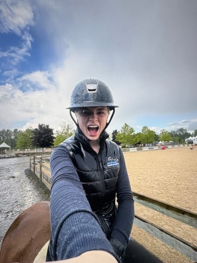 The image is a low-angle selfie taken outdoors at an equestrian facility. The primary subject is a young person, likely female, with short blonde hair, seated on a partially visible brown and white horse. The individual wears a dark, water-droplet-covered equestrian helmet and a black, shiny puffer vest over a dark, long-sleeved ribbed shirt. A logo reading "BCETT Equine" is visible on the left chest of the vest. The subject's mouth is wide open in an expressive, shouting posture.
The immediate environment features a light tan sand arena on the right and a dark, wet, muddy dirt path or another arena on the left, both separated by a light-colored, three-rail wooden fence. In the background, deciduous trees are prominent, with several white tents or structures visible beyond the arenas. The sky above is partly cloudy, exhibiting both blue sky and grey cloud formations, consistent with recent or ongoing precipitation, as evidenced by water droplets on the helmet and the muddy ground.
Based on the "BCETT Equine" logo, the location is within British Columbia, Canada. The specific city within a generic "Electoral Area A" cannot be determined from the visual evidence, as "Electoral Area A" typically refers to unorganized or rural areas within regional districts rather than a specific city.