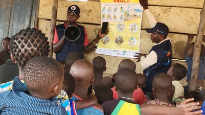 In Bunia, Democratic Republic of the Congo, a group of children is gathered outdoors, attentively listening to two men. One man, wearing a dark vest over a red shirt and a cap, holds a megaphone and points to a poster on a wooden wall. The poster, with French text and illustrations, appears to be an informational campaign about Ebola. The second man, also wearing a vest with a logo and a cap, stands beside the poster, gesturing towards it. The environment is rustic, with visible wooden structures and natural lighting. The children are of varying ages, some with their backs to the camera, and they are positioned in front of the men, indicating they are the audience. The scene suggests an educational or awareness-raising event.