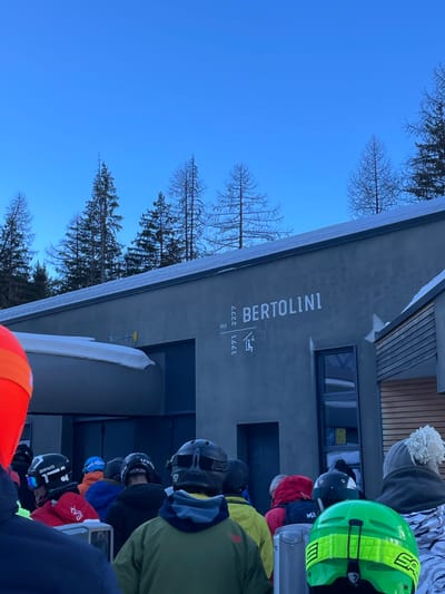 The image, oriented correctly, depicts a winter scene at a ski resort in Courmayeur, Italy, likely near a mountain lodge or a ski lift entrance. A modern-style building with a dark grey facade dominates the right side of the frame. Vertical white lettering on the building clearly displays "BERTOLINI," beneath which are the numbers "1771" and "2277," followed by "mt," suggesting altitudes in meters. A small graphic symbol resembling a person ascending steps is situated above these numbers.

In front of the building, a line of skiers or snowboarders, clad in winter apparel including colorful helmets (notably bright green, black, and orange), wait in what appears to be a queue. Their faces are largely obscured by helmets or turned away. Snow covers the roofline of the building and a curved structure, possibly a barrier for the queue, in the foreground. The background features a clear, bright blue sky and a mix of bare deciduous trees and dark evergreen conifers, indicative of a cold, clear day in the mountains. The overall impression is a bustling yet serene moment at a ski facility.