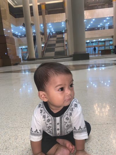 A baby is depicted sitting on a light-colored, speckled floor within a spacious indoor environment. The baby, with dark hair and eyes, is wearing a white long-sleeved top featuring black and grey geometric patterns, paired with black shorts or pants. The child is looking upwards and slightly to the right, with a calm and curious expression.

The setting appears to be a large public building, possibly a mall, hotel lobby, or convention center, consistent with a location like Samarinda, Indonesia. The background showcases multiple levels of the building, characterized by polished floors, reflective surfaces, pillars, and railings. Numerous artificial white lights are visible throughout the distant background, casting bright reflections on the floor, suggesting a well-lit indoor space, potentially during the evening or night. The overall atmosphere is clean, modern, and expansive. There is no specific activity beyond the baby observing its surroundings, and no visible text.