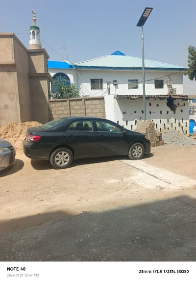 The image displays an outdoor, sunlit street scene in Kofar Ruwa, Nigeria, under a clear sky. The ground consists predominantly of unpaved, light brown earth, with a partial concrete-paved section in the immediate foreground. On the left, an unrendered light brown building is partially visible, adjacent to a large pile of light-colored sand. Above this structure, a white minaret with a green dome and a golden crescent moon finial is prominent. A black sedan is parked facing right on the dirt road in the midground, with the front bumper and fender of a lighter-colored vehicle partially visible on the far left, obscuring part of the black sedan's front. Behind the black sedan, a low concrete block wall runs horizontally, separating the unpaved area from a two-story white building. This building features a light blue hip roof, multiple rectangular windows, and a distinctive arched window on the first floor. An external air conditioning unit is mounted on its wall. To the right of the black sedan, a stack of concrete blocks and a small pile of gravel are present on the ground. A tall metal pole equipped with a solar-powered LED street light fixture stands upright to the right of the black sedan, positioned in front of the white building. Overhead wires are visible crisscrossing in the upper left portion of the frame. No individuals are present in the scene, and no explicit actions or interactions are depicted.