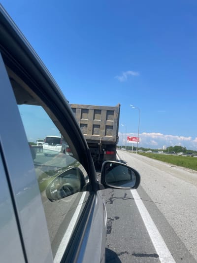 The image shows a view from inside a vehicle, looking out the side window towards a bright blue sky with a few wispy clouds. The foreground is dominated by the interior of the car, with a portion of the door frame and window visible. The side mirror is also in frame, reflecting the road ahead and other vehicles.
The main subject is a large, empty dump truck directly in front of the camera vehicle, in what appears to be a traffic jam on a highway. Beyond the dump truck, other vehicles can be seen, including a white minivan and a white truck further down the road. The highway has a clear white lane marking and is bordered by a grassy verge. In the distance, there are trees and some buildings, suggesting an urban or semi-urban environment. A billboard with Russian text and a red graphic is visible on the right side of the road. The overall impression is a sunny day on a busy road, possibly with a traffic delay. The location is identified as Solovej-Kljuch, Russia, suggesting this is a view of a highway or major road within or near that locality.