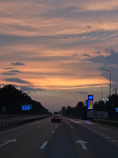 The image depicts a highway scene at dusk near Velyka Dymerka, Ukraine. The sky is filled with dramatic clouds, tinged with orange and pink from the setting sun. A multi-lane highway stretches into the distance, with a few cars visible. On the right side of the highway, a gas station sign with illuminated prices stands tall, and streetlights line the road. Trees form a dark silhouette along the left side of the highway. The road is marked with white lines, indicating lanes and directional arrows. The overall atmosphere is serene and somewhat melancholic, characteristic of a sunset on a highway.