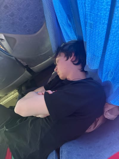 A person is sleeping on a bus. The person has dark hair and is wearing a black shirt. The bus has blue seats and curtains. The location is Челва, Russia.
