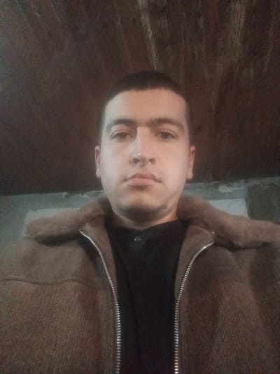 A low-light portrait depicts an adult male individual, centered in an indoor setting, stated to be Kasan, Uzbekistan. The subject has short, dark hair, dark eyebrows, and a neutral expression, gazing directly forward into the camera. He is dressed in a dark, possibly black, collared shirt underneath a brown jacket. The jacket features a distinct fur-like collar in a matching brown tone and a visible silver-colored zipper running down the front. The background above the subject's head consists of dark wooden planks or panels. To the sides of the subject's shoulders, the background is a darker, indistinct wall, with some lighter, possibly peeled, areas discernible on the upper left side of the frame. The scene is dimly illuminated, contributing to the dark overall exposure of the image.