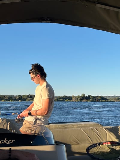 On a boat on the Zambezi River in Victoria Falls, Zimbabwe, a man wearing sunglasses, a t-shirt, and shorts is fishing. The river is lined with green trees and bushes. The sky is blue.