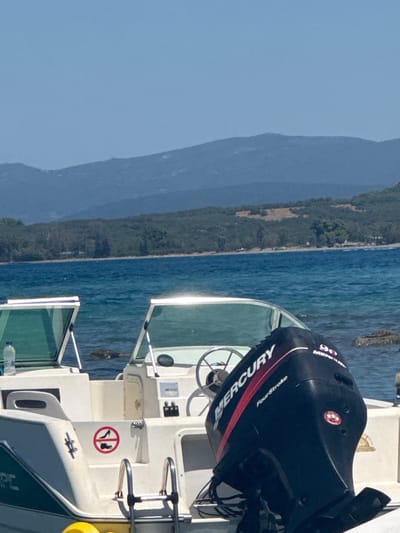 The image shows a close-up of a white boat with a black outboard motor, branded "Mercury FourStroke 90," docked in the clear blue waters of Lichada, Greece. The boat features a steering wheel and a red circular decal with a shoe and a high heel crossed out, indicating a no-shoes policy. In the background, a lush green coastline with hills and mountains stretches towards a hazy blue sky. The water is calm, with gentle ripples and visible rocks and seabed near the boat. The sun reflects brightly off the water and the boat's windshield.
