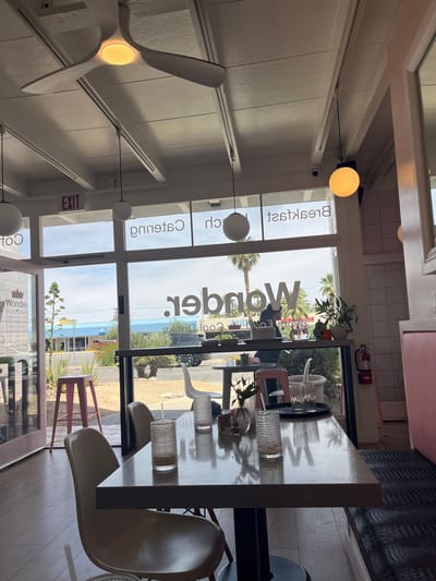 Here's a forensic description of the image:
**Scene:** Interior of a dining establishment, possibly a cafe or restaurant, located in Palm Desert, United States of America.
**Environment:** The space is brightly lit, appearing to have large windows that offer an exterior view. There is an exit sign visible.
**Objects:**
* Tables and chairs are present. The chairs are white in color with a distinctive modern design.
* Various objects (glassware, decorative items) are on the table.
* A ceiling fan is visible, with a yellow light.
* A fire extinguisher is mounted on the wall near the top of the window.
**Subjects:**
* The windows feature text indicating the name "Wonder," along with phrases like "Cool Drinks" and menu or business hours.
* Outside of the windows, there are palm trees, buildings, and cars visible, suggesting an urban or suburban location.
**Actions/Interactions:** No people are actively present or engaged in any visible action.