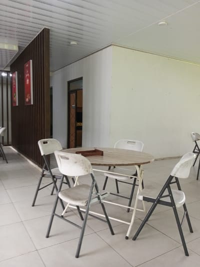 The image depicts an unoccupied indoor common area or dining space located in Pachmarhi, India. The floor is covered with light-colored square tiles. In the foreground and center, a round table with a light-colored, possibly marble-patterned, top and white metal legs is surrounded by four folding chairs. These chairs feature white plastic seats and backs with dark grey metal frames, exhibiting visible signs of wear and discoloration. A rectangular wooden tray rests on the table's surface. To the left, a dark brown wooden slatted room divider stands vertically, with two framed posters displaying red backgrounds and white graphic elements mounted on it. Further to the left, additional chairs are partially visible. In the background, a doorway with a dark frame opens into an interior room. The walls are painted a plain off-white, and the ceiling consists of white horizontal paneling with several recessed circular light fixtures or smoke detectors. No people, actions, or interactions are present in the scene.
