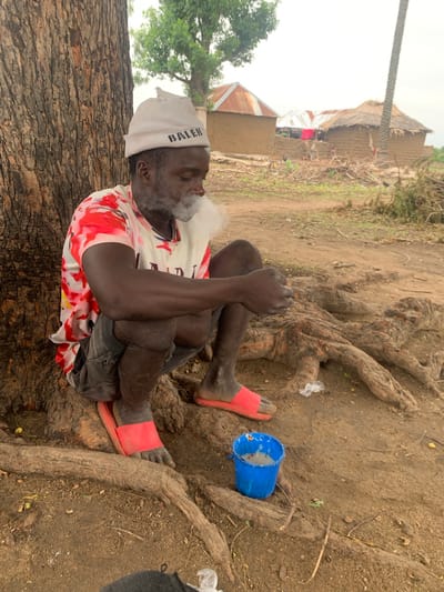 A Black man, wearing a grey knitted beanie with "BALEN" text, a red and white tie-dye t-shirt, and dark shorts, is crouched by the base of a large tree. He is smoking and exhaling a cloud of white smoke. His right hand is near his mouth, and his left hand rests on his knee. He is wearing red slide sandals. Beside him on the ground is a small blue bucket containing a light-colored substance, possibly food or liquid. The ground is covered in dirt and exposed tree roots. In the background, there are several mud-brick structures with corrugated metal and thatched roofs, indicative of a rural setting. The sky is overcast. This scene is located in Dull, Nigeria.
