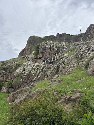 The image shows a rocky mountain landscape under an overcast sky. The mountain is covered with varying amounts of grass and sparse vegetation. A power line stretches across the sky. The image was taken in Каменномостское сельское поселение, Russia.