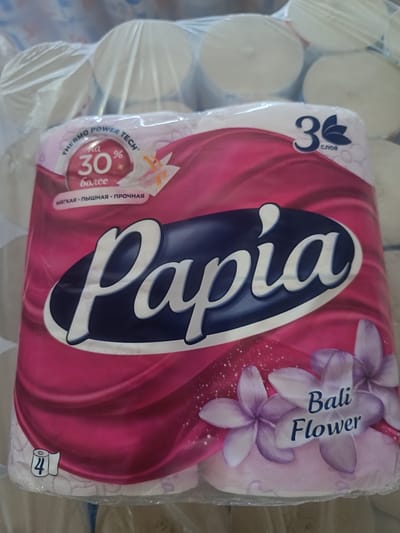 This is a close-up, slightly angled, eye-level indoor photograph of a multi-pack of toilet paper rolls. The product packaging features a prominent "Papia" logo in a dark blue oval with white lettering. The packaging is predominantly pink with swirling patterns and floral graphics, specifically depicting purple and white flowers that appear to be Plumeria or Frangipani. Text on the packaging includes "THERMO POWER TECH", "на 30% более", "мягкая • пышная • прочная", "3 слоя", and "Bali Flower". A small graphic of a toilet roll with the number "4" is visible in the lower left corner. The toilet paper rolls are wrapped in clear plastic, and the background shows more stacked rolls of toilet paper, also wrapped in plastic. The lighting appears to be artificial, suggesting an indoor environment such as a retail store or storage area. There are no people visible in the image. The scene is a product display.