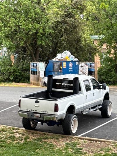 A white Ford Super Duty pickup truck is parked at an angle on an asphalt parking lot next to a grassy patch. The truck has oversized tires and exhaust stacks. A blue dumpster overflowing with garbage is behind the truck. Trees surround the parking area. The license plate is 4FT2134. The location is in Alexandria, United States of America.