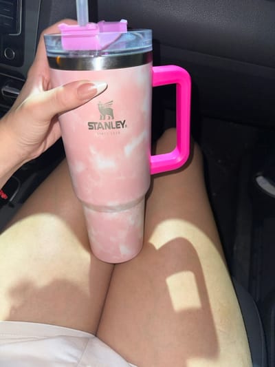 A pink Stanley tumbler with a pink handle sits on a woman's lap. The tumbler is light pink with a cloudy, marble-like pattern, and the word "STANLEY" is printed on its side. The tumbler has a clear lid with a pink straw and a pink mechanism. The woman holds the tumbler with her left hand, showing a manicured nail. The background is the dark interior of a car.