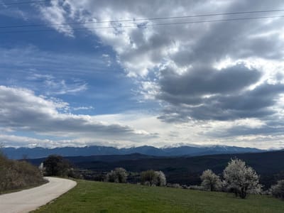 A landscape scene in Ochusha, Bulgaria. A curved dirt road leads into the distance, framed by a green grassy hill on the right and trees on the left. Several flowering trees with white blossoms dot the hillside. In the background, a dark, forested range of hills leads to a mountain range capped with snow. Above the mountains, the sky is filled with a mixture of blue sky and heavy gray clouds. Power lines are visible across the top of the frame.