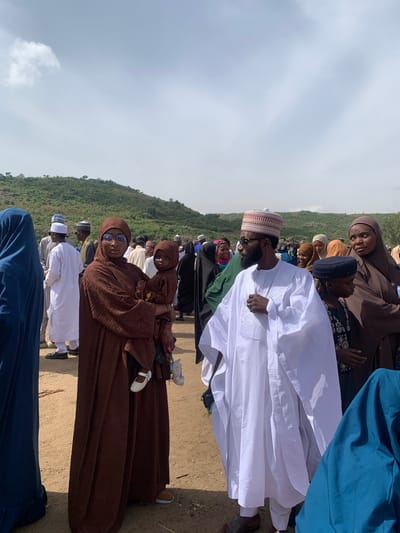 People stand outdoors on a dirt surface near a hill. The sky is cloudy. Many people wear traditional Muslim clothing and head coverings. Some wear glasses. One person holds a child. Naraguta Hausawa, Nigeria is not visible.