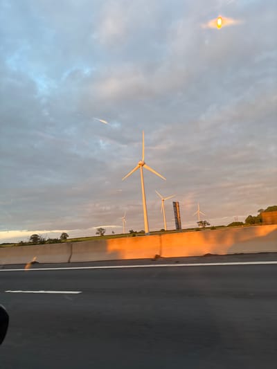The image, captured from a moving vehicle, is oriented vertically (portrait) and depicts a view from a highway near Roade, United Kingdom, featuring multiple wind turbines under a partly cloudy sky during the golden hour.

Along the right side of the frame, a dark asphalt highway extends vertically, with a distinct white dashed line marking lanes. Adjacent to the highway is a tall concrete barrier or wall, which prominently reflects a warm, orange-yellow glow from the low sun, creating a striking visual contrast with the cooler, bluer and grey tones of the sky.

Towards the middle and left of the frame, several wind turbines are visible. The most prominent one is in the mid-ground, extending leftwards with its long, white blades. Further back and slightly to the right, additional wind turbines can be seen, some partially obscured by the distant landscape. The sky above is a mosaic of light blue, grey, and soft orange clouds, illuminated by the low-angle sunlight, indicating either sunrise or sunset. Beyond the turbines and barrier, a stretch of green land with scattered trees and vegetation can be observed. The overall scene conveys a sense of calm and industrial scale against a natural backdrop. No discernible text is visible on any objects in the image.