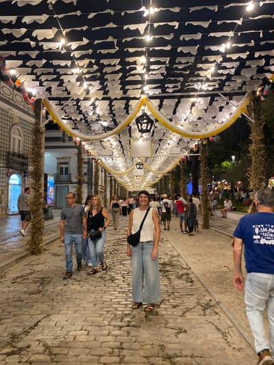 A nighttime street scene in Tavira, Portugal, features a cobblestone path lined with buildings and trees, all under a canopy of white paper flags and yellow and white garlands with string lights. In the foreground, a woman with short, dark hair and a white top and wide-leg jeans smiles at the camera. She wears a black crossbody bag and dark sandals. Behind her, a man in a blue t-shirt with the text "SÃO JOÃO TAVIRA" walks away from the camera. Several other people are visible walking along the street, some in pairs and groups, and some are seated on a low wall in the background. The street is illuminated by the overhead lights and lights from the surrounding buildings, creating a festive atmosphere.