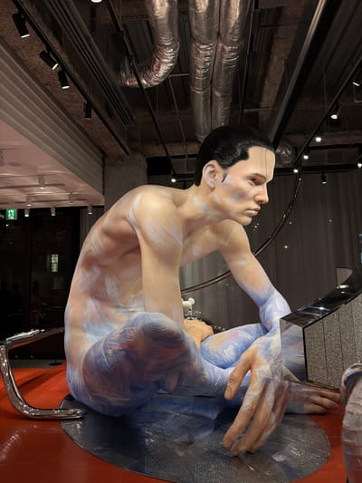 The image shows a life-sized statue of a person, painted with blue, white, and pink brushstrokes, seated in a cross-legged position. The statue is located inside a commercial establishment, likely a store, as indicated by the presence of track lighting and exposed industrial ductwork on the ceiling. The background features a large window, through which a blurred view of the street and other buildings can be seen. The statue is positioned on a red circular platform, with a metallic chrome-colored structure to its left. A black, textured, rectangular object is placed to the statue's right. The overall impression is that of an art installation or display. The store is identified as "DOVER STREET MARKET GINZA", located in Chuo, Japan.