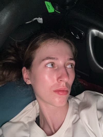 The image shows a close-up, overhead shot of a young Caucasian woman lying down, likely in a car, based on the dark, textured material resembling upholstery and a chrome-colored handle visible. She has light brown hair pulled back, and her head is tilted to the right, with her eyes looking off to the side. Her skin appears flushed and slightly shiny, and she is wearing a light-colored sweatshirt. In the background, there is a green object that might be a marker or highlighter, and a crumpled piece of white paper on the dark surface. The overall lighting is dim, suggesting an indoor or nighttime setting. The image does not contain any elements that would allow for identification of a specific location within Lasva, Russia.