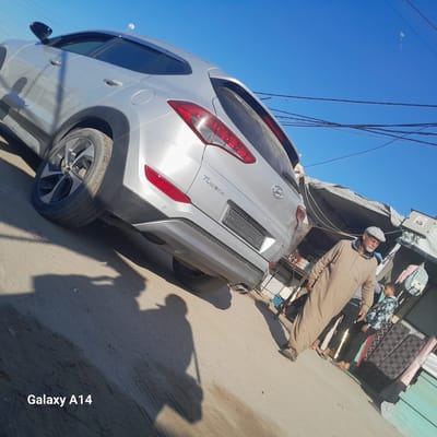 The image, taken in Al-Mawasi, shows a silver Hyundai Tucson parked on a paved surface near a street. An older man wearing a long beige garment and a cap is standing next to the car, seemingly engaged in a conversation with a younger boy who is wearing a patterned jacket and dark pants. The background includes a makeshift shelter or shop, with various items and goods visible. Power lines run overhead against a clear blue sky. The shadow of the photographer is prominent in the foreground.
