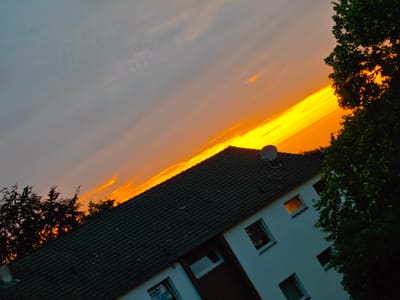 The image depicts a diagonal view of a residential building against a dramatic sunset sky. The building is white with a dark tiled roof and several windows, some of which appear lit. A satellite dish is visible on the roof. Lush green trees frame the right side of the image and are silhouetted against the bright orange and yellow hues of the sunset on the left. The sky is a gradient of grey and orange, with streaks of bright yellow indicating intense sunlight. The photograph appears to have been taken in Georgsmarienhütte, Germany, likely during an evening hour. There are no people visible in the image. The composition is skewed, giving the impression of a tilted perspective.