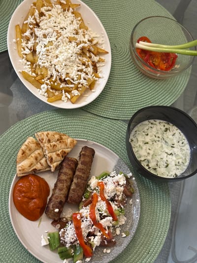 A top-down view depicts a meal spread across a light-colored surface, likely a table, over two light green, circular woven placemats. The specific city of Naples, United States, cannot be determined from the visual information provided.
On the upper placemat, a white oval plate holds a serving of golden-brown French fries, generously topped with crumbled white cheese, possibly feta. Adjacent to this, a small, clear glass bowl contains sliced red peppers and two stalks of green onion.
On the lower placemat, a round, grey patterned plate presents three pieces of grilled flatbread, two elongated, grilled meat rolls (e.g., cevapi), and a dollop of thick orange-red sauce. A salad occupies the remainder of the plate, consisting of diced cucumbers, tomatoes, and strips of red bell pepper, also heavily garnished with crumbled white cheese. To the right of this plate, a black bowl holds a creamy white sauce containing diced cucumbers and green herbs, consistent with tzatziki. No individuals are visible in the scene, and no specific actions or interactions are depicted beyond the presentation of the prepared meal.
