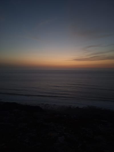 The image depicts a tranquil seascape at dusk or dawn, likely near Ahrara, Morocco. The sky transitions from a deep blue at the top to a soft orange and yellow glow near the horizon, indicating the sun is either rising or setting. Wispy clouds are scattered across the upper portion of the sky. Below the sky, a vast expanse of the ocean stretches out, appearing somewhat dark and calm with subtle waves. The foreground is dominated by a dark, rocky shoreline or cliffside, rendered almost entirely in shadow, with only faint textures visible. The overall atmosphere is serene and somewhat moody, characteristic of twilight hours over the Atlantic coast.