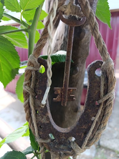 A rusty horseshoe is suspended by a thick rope, from which an old, rusty key hangs in its center. The horseshoe is attached to a wooden post, which is partially visible. Green leaves of a plant are in the foreground and background. The background appears to be a blurred outdoor setting with a red structure. The overall scene suggests an outdoor decoration or symbolic arrangement, possibly in a garden or rural area. No people are visible in the image. The location in Filippenkov, Russia cannot be determined from the image itself.