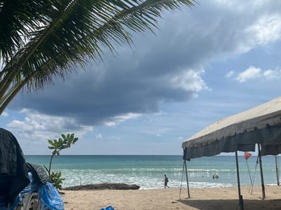 A wide-angle, eye-level outdoor shot captures a beach scene in Ban Bang Thao, Thailand. In the foreground, a large, weathered tent with a beige canopy is pitched on the sand, supported by dark poles. To the left of the tent, a pile of white lounge chairs is covered with blue plastic sheeting, and a black tarp is draped over some of the chairs. A small tree with vibrant green leaves grows in the sand between the foreground objects and the ocean.
The middle ground features a sandy beach with gentle waves lapping the shore. A single person is walking along the edge of the water, and several other individuals are visible wading or swimming in the shallow ocean. A weathered piece of driftwood lies on the sand near the water's edge.
The background consists of the turquoise ocean stretching to the horizon, meeting a sky filled with dramatic, dark storm clouds juxtaposed with patches of bright blue sky and scattered white clouds. The fronds of a palm tree extend from the upper left corner of the frame, partially obscuring the sky.