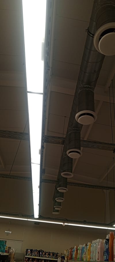 The image depicts the interior ceiling of a commercial establishment, likely a supermarket, in Zhirnovsk, Russia. Dominating the upper portion of the frame are two long, bright fluorescent light panels, characteristic of commercial lighting. Below these, a series of cylindrical air ducts, made of corrugated metal, run diagonally across the scene from right to left. Attached to these ducts are circular white vents, spaced at regular intervals, which appear to be air conditioning or ventilation outlets. The ceiling itself is constructed of beige-colored panels. Below the ceiling, a metal shelving unit filled with various products is visible at the bottom of the frame. The products appear to be packaged goods, possibly cleaning supplies or toiletries, given the diverse colors and shapes of the bottles. A sign that reads "ВЫХОД" (EXIT in Russian) is visible above a doorway on the left side of the image. The overall impression is one of a functional, indoor retail space.
