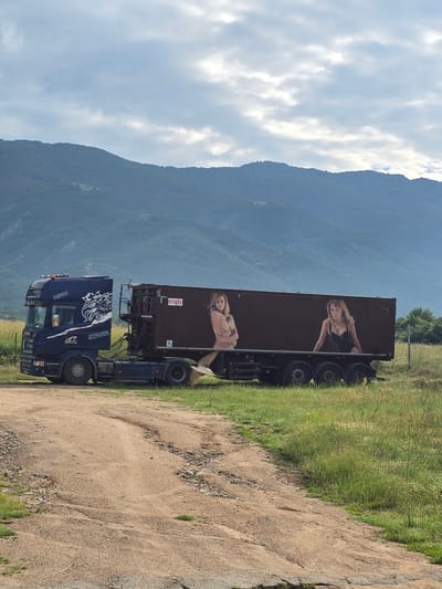 A blue Scania R580 truck is parked on a dirt path in a grassy field in Karnare, Bulgaria. The truck's cab features a white graphic of an eagle-like creature and the word "SCANIA" on its side. Connected to the truck is a dark brown trailer, which displays two large painted images of women. The woman on the left is depicted nude from the waist up, with her arms crossed, while the woman on the right wears a black low-cut top. A red rectangular label with the white numbers "91553" is visible on the upper left side of the trailer. The ground consists of dry, light brown dirt with tire tracks and patches of green grass and weeds. In the background, prominent dark green, tree-covered mountains rise under a partly cloudy, light blue sky. A small, light-colored bird is captured in flight near the lower right side of the trailer.