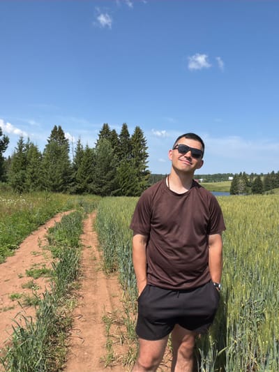 A medium shot captures a man standing on a dirt path in a rural landscape. He is wearing sunglasses, a brown t-shirt, and black shorts. The path runs through a field of tall green grass, with a dense forest of evergreen trees visible in the background. The sky is a clear blue with a few scattered white clouds. The location is identified as Podshivalovo, Russia.