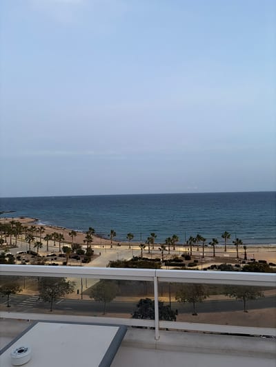 The image presents an elevated view of the coastline in Villajoyosa/la Vila Joiosa, Spain, observed from a balcony. In the immediate foreground, a white rectangular table with a gray perimeter is partially visible on the left, holding a white cylindrical ashtray. A white railing with transparent glass or plastic panels spans horizontally across the lower portion of the frame. Beyond the railing, a paved road with a visible pedestrian crossing on the left is present, accompanied by small trees and illuminated streetlights. Further back, a wide, level promenade constructed of concrete or paving stones runs parallel to the water's edge, densely lined with numerous equidistant palm trees. This promenade gives way to a sandy beach. The Mediterranean Sea occupies the midground and background, characterized by calm, dark blue-green water extending to a faint horizon. The sky is a uniform light blue-gray, indicative of an overcast condition or twilight. No human subjects are discernible within the depicted environment.