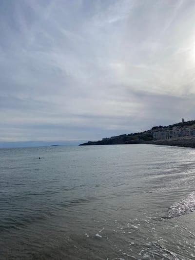This image captures a coastal scene in the city of Sète, France, under an overcast sky. The sky is dominated by layers of light grey clouds, with a concentrated bright area in the upper right quadrant indicating a hidden sun. The sea occupies the lower two-thirds of the frame, displaying a calm, rippled surface with a grey-blue coloration. In the mid-ground, a single individual is visible swimming, marked by a distinct small red object, likely a swim cap or safety buoy. Along the lower right edge of the image, small waves gently break on the shoreline, producing white foam.
The right side of the photograph features a prominent landmass rising from the water, a hill densely populated with numerous multi-story buildings, appearing to be residential or commercial structures. A slender, white, cylindrical tower, consistent with a lighthouse, is visible at the crest of this hill. The immediate coastline beneath the buildings transitions from rocky outcrops to a lighter-colored sandy or pebbly beach where the water meets the land. On the far left horizon, faint, low-lying landforms are discernible. The overall lighting is soft and diffused, characteristic of an overcast day.