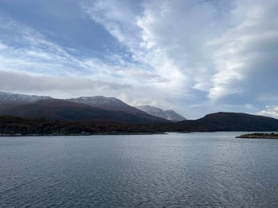 The image presents a wide body of water, possibly a lake or a sea inlet, reflecting the overcast sky. In the background, a chain of mountains is visible. Some of the peaks are capped with snow or ice. A band of dense forest lines the shore between the water and the base of the mountains. The sky is filled with clouds, ranging from thin cirrus formations to heavier, darker storm clouds. The overall impression is one of a remote, natural landscape. Based on the prompt, the location is Lapataia, Argentina. No people are visible in the image. The subject is nature. A small island to the right side of the frame. The time of day appears to be daytime.