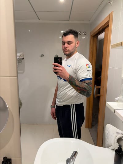 A male individual, appearing to be in his 20s or early 30s, with short dark hair and light stubble, is depicted taking a mirror selfie in a well-lit bathroom. He is observed from the chest up, with his lower body partially visible. He wears a white, short-sleeved athletic shirt featuring a blue Nike swoosh logo on the right chest area and a colorful LaLiga logo patch on the right sleeve. His left arm is covered by a detailed, colorful tattoo sleeve, likely depicting a dragon or similar motif. He is also wearing navy blue athletic sweatpants with three white stripes down the side of each leg and a metallic watch on his left wrist. The subject holds a black smartphone in his right hand, positioned to capture his reflection.
The background reveals a modern bathroom setting with white tiled walls and a white tiled floor. A white ceramic sink with a chrome faucet is visible in the lower foreground. The ceiling features square panels and recessed lighting. Chrome towel hooks are mounted on the wall behind the subject. To the right, a wooden door frame, approximately light brown in color, borders a partially open wooden door, revealing a dimly lit interior. A small wooden shelf is mounted on the wall above the door frame. A white folded towel is draped over a surface or rack on the far right, and a black pump-action dispenser bottle is situated on a counter next to the sink. The specific location within Villajoyosa/la Vila Joiosa, Spain, cannot be determined from the visual evidence, but the environment is consistent with a hotel or modern apartment bathroom.