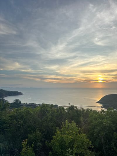The image shows a vertical view of Baan Wang Ta Kien, Thailand, at sunset. The sky, filled with wispy clouds, transitions from a bright, sunlit area near the horizon to a darker shade of gray. The sun's golden light reflects on the calm water surface. Green foliage occupies the right side of the image, bordering the sea. In the distance, land masses appear along the horizon.