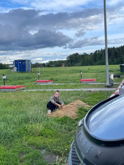 A photo taken outdoors in Бавыкино, Russia, shows a boy crouching next to a dirt pile with a green grassy field behind him. Behind the boy, several red and gray markers stand in the field. Further back, a blue metal structure, identified as equipment building ПУ-Р-4х4В, is situated behind a chain-link fence. In the top right, there's part of a black object with the word "WERK" visible. The sky above is filled with cloudy formations.