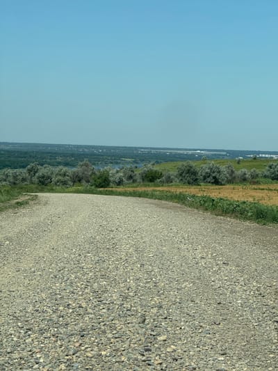 The image captures a scenic view from a gravel road in Sukhoi, Russia. The road, covered in small, rounded stones, curves gently towards the right of the frame, leading the viewer's eye into the landscape. To the left and ahead, a verdant valley unfolds, dotted with numerous trees and shrubs. A body of water, possibly a river or a lake, is visible in the mid-distance, reflecting the clear, pale blue sky. Beyond the valley, a distant treeline marks the horizon, with hints of buildings and structures visible, suggesting a settlement or town in the far background. The sky is vast and cloudless, indicating a sunny day. The overall impression is one of a tranquil, rural landscape.