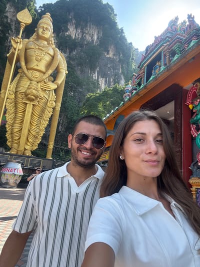 A portrait-oriented photograph captures a man and a woman posing together in front of a monumental golden statue and a vibrant temple structure, set against a backdrop of a lush, rocky cliff face. The scene is brightly lit by daylight, suggesting clear weather.

In the foreground, slightly to the right, are the two individuals. The woman, with long brown hair, is wearing a white collared top and a small stud earring, smiling subtly towards the camera. Beside her, the man, with a short beard and dark sunglasses, smiles broadly. He is wearing a white polo shirt with horizontal dark grey stripes.

To their left stands a towering, intricately detailed golden statue of a deity, likely Lord Murugan, holding a spear-like object. This prominent statue suggests the location is a significant religious or cultural site. Behind the subjects and the statue, parts of a colorful temple facade are visible, adorned with traditional, ornate carvings in shades of green, blue, red, and orange.

The background features a steep, verdant mountain or cliff face, covered with trees and foliage. The overall setting, including the golden statue and temple architecture, strongly points to Batu Caves in Selayang Municipal Council, Malaysia. Both individuals appear happy and relaxed, enjoying their visit. Partially visible text "DONATION" can be seen on a container or sign near the man's lower right, and some indistinct text in Tamil script is on a sign behind the woman.