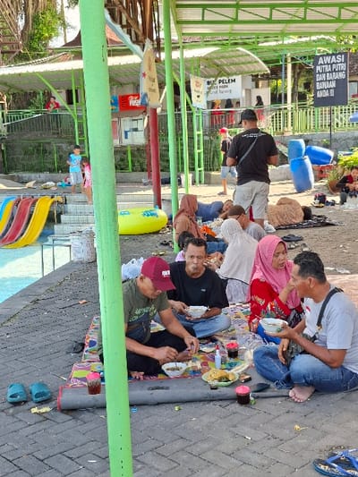 A group of people are gathered at an outdoor eating area in the city of Kecamatan Tulung, Indonesia, located at latitude -7.592 and longitude 110.65. They are sitting at a picnic table, eating and talking. There is a green pole in the foreground and a partially visible swimming pool in the background. Several people are sitting at the table, with one man and one woman actively eating. The man is wearing a red cap and a green shirt, while the woman is wearing a white headscarf and a red shirt. Other people are sitting at the table, observing the scene or eating as well. The people are sitting on a colorful mat. The ground is made of gray concrete. There are some signs on the walls, including a sign that says "AWASI PUTRA PUTRI DAN BARANG BAWAAN ANDA!!!". The scene is calm and relaxed. The sky is clear, and there are trees and bushes in the background. There are also some blue plastic barrels near the table.