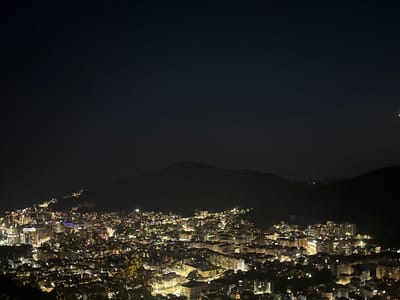 A nighttime aerial view captures the sprawling city of Košljun, Montenegro, nestled at the base of dark, silhouetted mountains. The city is illuminated by a dense network of artificial lights, creating a vibrant tapestry of yellow, white, and occasional blue glows emanating from buildings and streetlights. The lights are most concentrated in the lower and central portions of the image, extending up the slopes of the hills. The sky above is a deep, dark blue, with a few faint stars visible, and a sliver of the moon is present on the far right edge of the frame. The foreground is dominated by the dark, out-of-focus foliage of trees and shrubs, suggesting the viewpoint is from a higher elevation. The overall impression is of a lively, populated urban center under a vast, clear night sky.