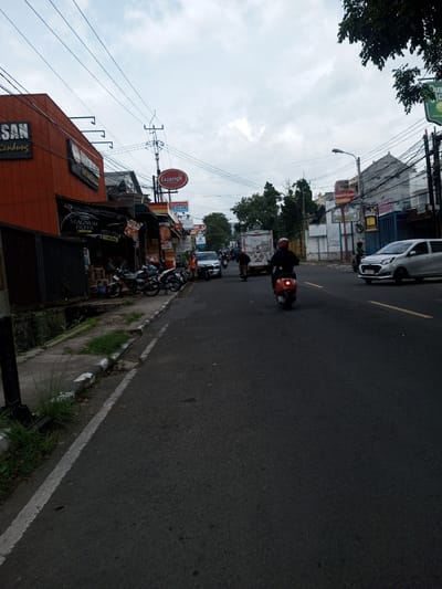 The image depicts an urban street scene, identified as being in the city of Khajuripada, India, under an overcast sky. The two-lane asphalt road appears dark and potentially damp, featuring a faded white solid line delineating the left shoulder and faded yellow double solid lines visible further in the distance.
On the left side of the street, a concrete curb is overgrown with green grass and weeds, bordering a dark metal fence. Behind the fence, a prominent orange building displays a dark sign with "SAN Bandung" in white text. Adjacent to this, a storefront features a dark awning and a sign reading "MAGNUM FILTER." Further along, additional commercial establishments include a building with a red oval 'Lazatto' sign and a blue 'APOTEK' sign. Multiple motorcycles are parked on the sidewalk/shoulder in this area. A silver SUV is also parked, with a person wearing an orange vest standing nearby.
In the foreground, a person wearing a dark helmet and dark clothing operates a red scooter, moving away from the camera, positioned slightly to the right of the road's center. Other motorcyclists and a white truck are observed moving in the same direction further down the street. A white compact car is visible in the right lane, heading towards the camera.
The right side of the street is lined with multi-story buildings of varying light colors, some appearing residential or mixed-use. A light blue container or modular structure is visible among them. Numerous utility poles with a dense network of overhead wires are present on both sides, with a street lamp visible on a pole on the right.