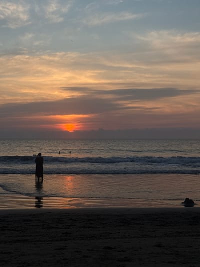A sideways view captures a beach at sunset. The sky transitions from light blue to fiery orange, reflected in the ocean waves. Silhouettes of people dot the shoreline, their forms stark against the shimmering water. The sand stretches into darkness on the right, contrasting with the vibrant hues of the sky and sea.