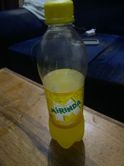 A clear plastic bottle of Mirinda pineapple-flavored soda lies horizontally on a wooden surface in Jama A, Nigeria. The bottle has a bright yellow screw cap and a yellow label wrapped around its middle section. The label features the brand name "MIRINDA" prominently in green letters with a white outline, and below it, the flavor "PINEAPPLE" is visible in smaller text. The bottle appears to be partially full of a yellow liquid, consistent with pineapple soda.
The wooden surface underneath the bottle shows a distinct grain pattern and appears worn, with several small dark specks and a lighter area suggesting indirect lighting. Behind the bottle, the background is dark, predominantly a deep blue or black, possibly a piece of fabric or an object. A small white object, resembling a cable or a charging adapter, is faintly visible in the dark background near the bottle's neck. The overall lighting suggests an indoor setting or a dimly lit area, as evidenced by the shadows cast by the bottle on the wooden surface. There are some faint, hard-to-read embossed markings on the clear plastic body of the bottle near the cap. No human subjects or distinct activities are visible in the image.