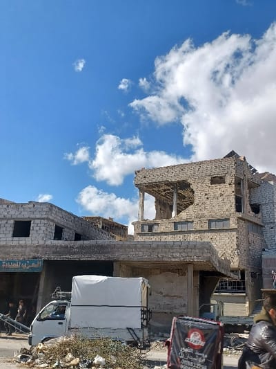 The image depicts an urban street scene in Al-Qadam Municipality, Syria, under a blue sky with scattered white clouds. The primary focus is on several multi-story buildings in the mid-ground, which exhibit extensive structural damage. The most prominent building on the right is constructed from light-colored concrete blocks and has significant portions of its upper floors and roof missing, revealing internal support structures and exposed rebar. Numerous impact marks or bullet holes are visible across its facade. An adjacent building to the left also shows signs of damage, and its ground floor features a storefront with an Arabic sign reading "العربي للمداواة" (Al-Arabi for Healing/Treatment).

In the mid-ground, a white pickup truck with a white canvas cover over its bed is parked, facing left. A male driver is visible through the windshield. A metal object, resembling a ladder or ramp, extends from the rear of the truck towards the left. In the foreground, a pile of assorted debris, including vegetative matter and trash, is accumulated on the street surface.

On the far right of the foreground, a person in a dark jacket is partially visible, obscured by a dark delivery bag or box. The bag features Arabic text identifying "مؤسسة الشهور فرع السيدان" (Al-Shuhour Foundation Al-Saydan Branch) and includes phone numbers (0118832294, 0996596956). One or two additional individuals are faintly visible beneath the awning of the building on the far left. The scene indicates an environment affected by conflict or severe disrepair, with ongoing commercial activity.
